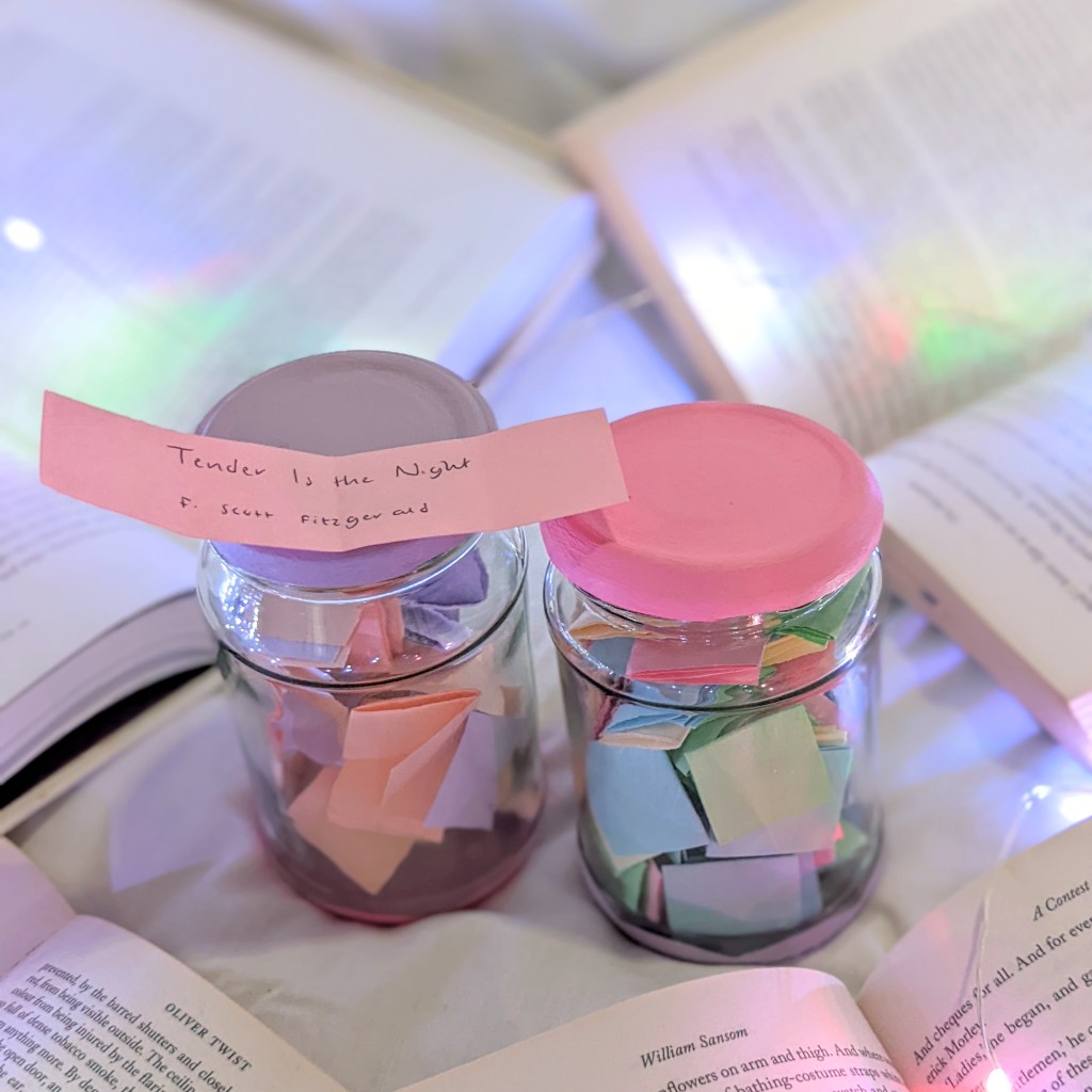 2 TBR jars on a bed surrounded by books. The jars are filled with folded up pieces of paper with the names and authors of books on them. One has been drawn and unfolded and says, 'Tender is the Night, F. Scott Fitzgerald'.